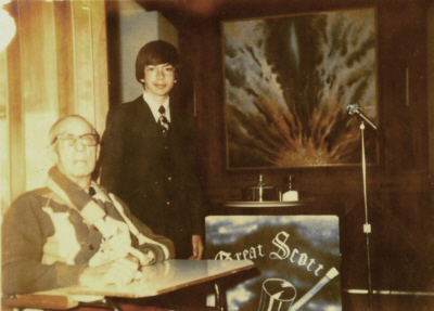 Two people in a room, one seated and one standing. There's a microphone and a podium with "Great Scot" inscription.