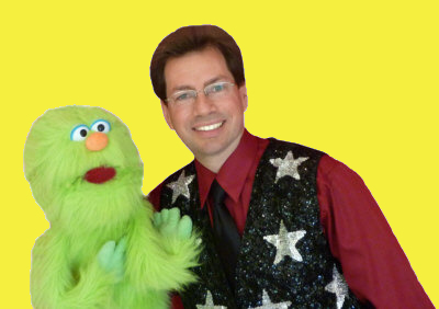 A person in a star-patterned vest poses with a green puppet against a yellow background.