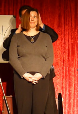 A woman stands in front of a man with her hands clasped, while the man holds her head, against a red curtain backdrop.