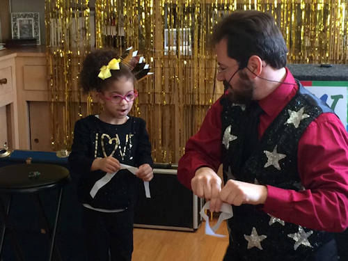 A magician performs a trick with a ribbon while a child with glasses and a headband observes, against a gold tinsel backdrop.