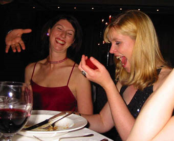 Two women sit at a dining table. One is laughing, while the other looks surprised, holding fruit. There's a hand reaching in.