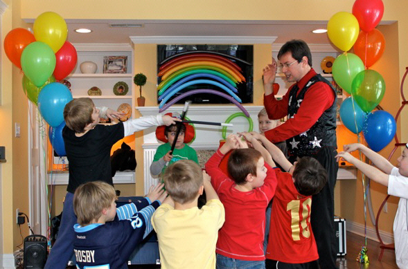 A magician performs tricks for a group of excited children at a party, with colorful balloons and a rainbow decoration in the background.