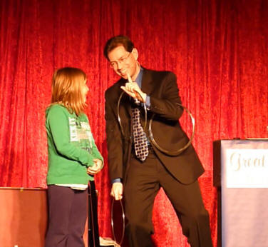 A man in a suit performs a ring trick on stage with a young girl in a green sweater, red curtain backdrop.