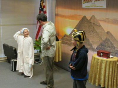 Two children wearing costumes stand with an adult in front of a backdrop featuring pyramids. An American flag is in the background.